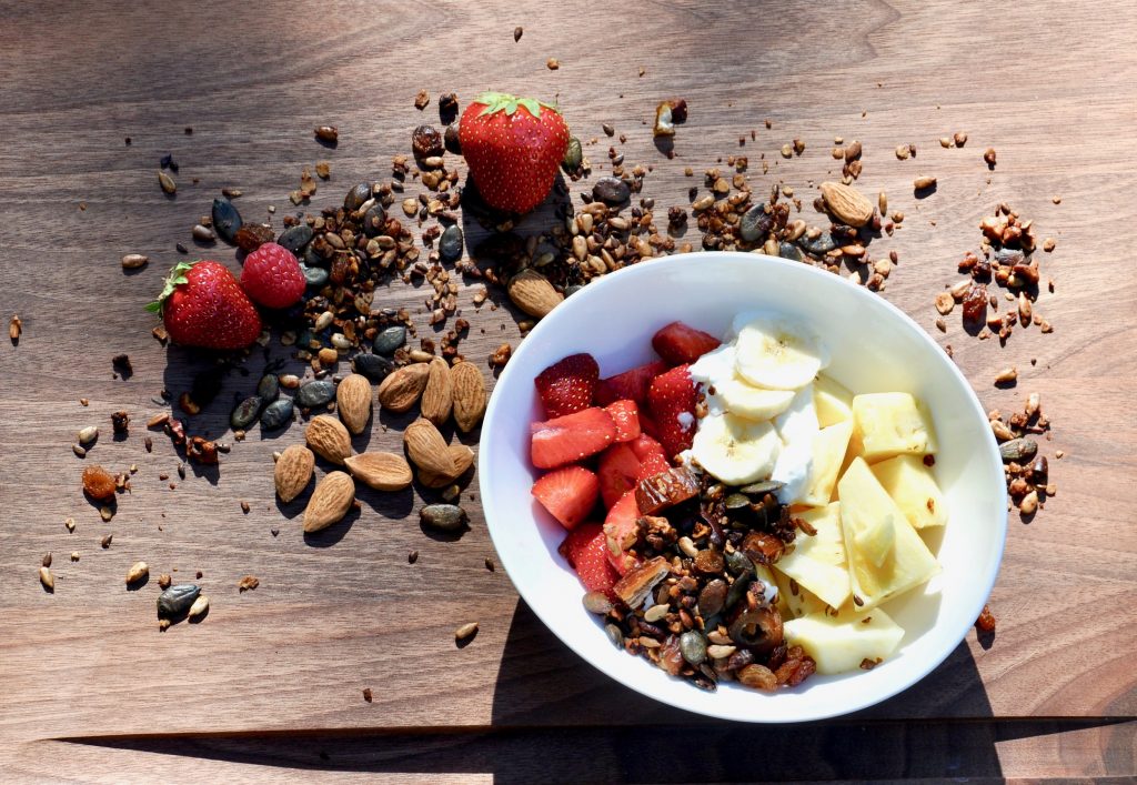 Eine Frühstücksschale mit Erdbeeren, Bananen, Äpfeln und Granola steht auf einer dunklen Holzunterlage - darum herum sind Erdbeeren, Nüsse und Granola verteilt