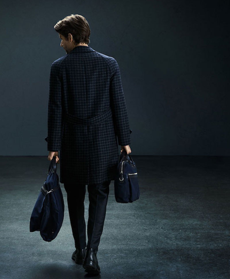 Ein elegant gekleideter Mann steht mit dem Rücken zum Betrachter vor einem dunklen Hintergrund und trägt zwei unterschiedliche Taschen von Mismo in den Händen
