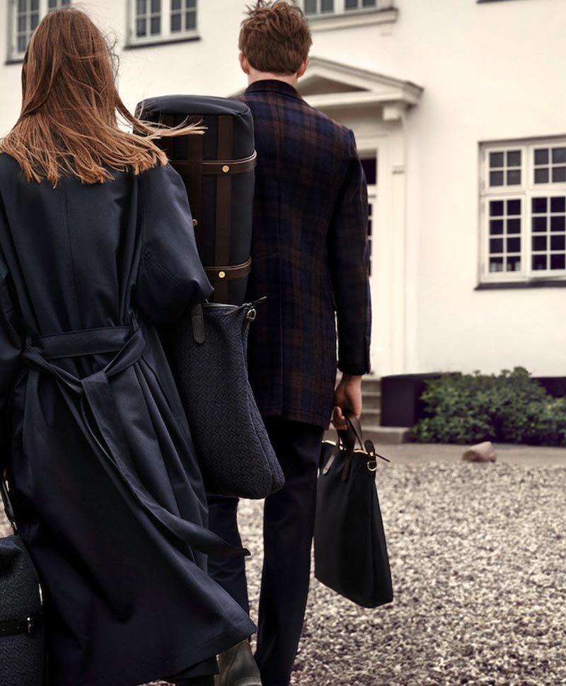 Zwei elegant gekleidete Personen sind im Begriff, ein Gebäude zu betreten - beide haben unterschiedliche Taschen der Marke Mismo bei sich