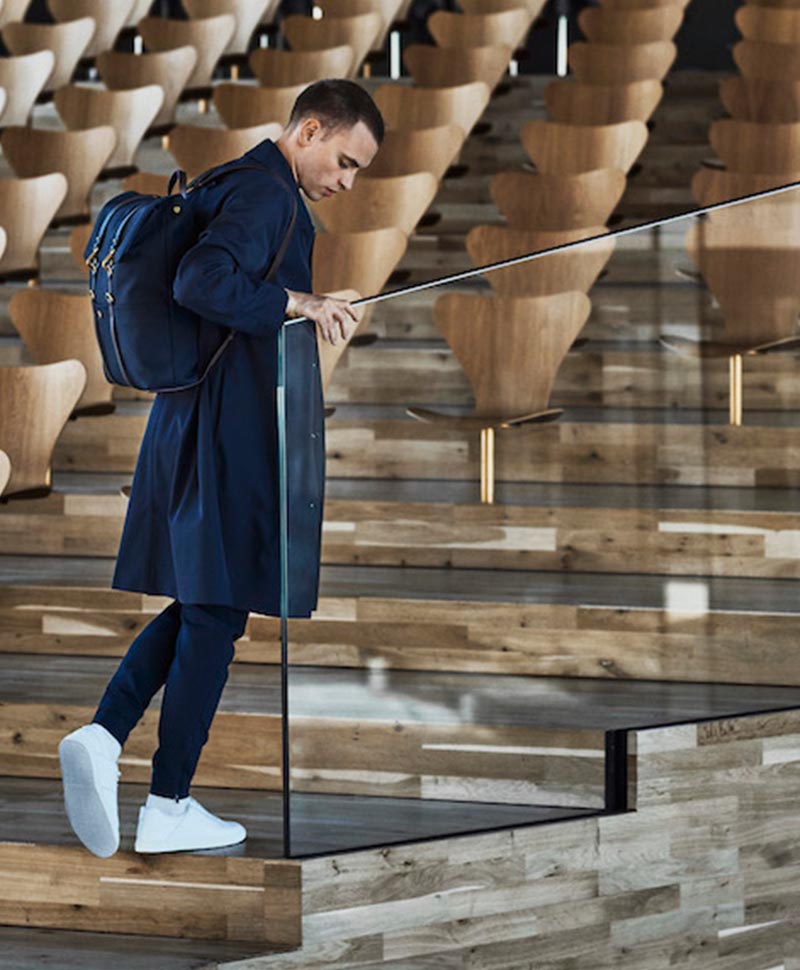 Ein elegant gekleideter Mann steigt die Stufen in einem Vorlesungssaal hinauf und trägt dabei den Rucksack "Backpack" von Mismo in der Farbe navy auf seinem Rücken
