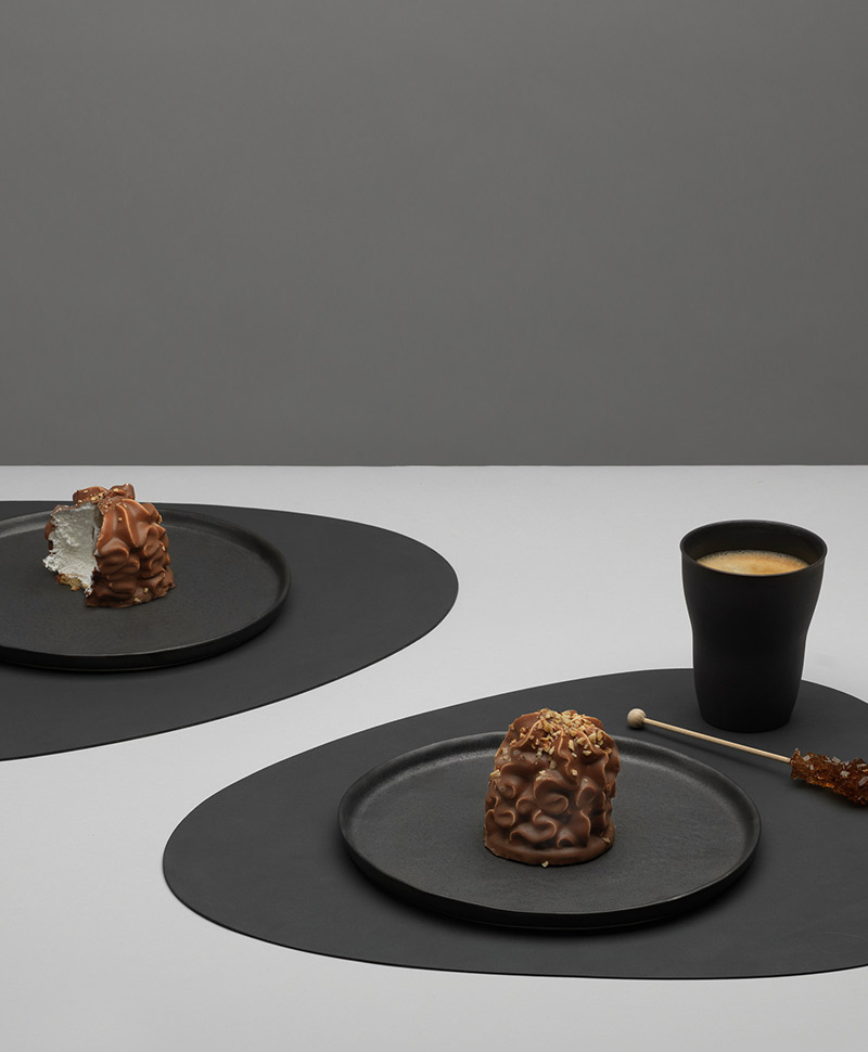 Moodfoto eines hellen Tisches, auf dem zwei schwarze Table Mats von Lind DNA, sowie darauf arrangierte Teller mit Dessert liegen