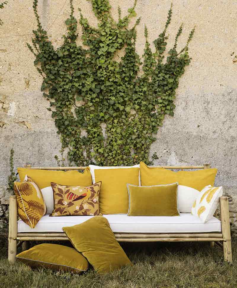frühlingshafte Kissen von Élitis in Ockertönen sind auf einem Sofa arrangiert, das auf einer Wiese vor einer rustikalen, efeubewachsenen Wand im Freien steht