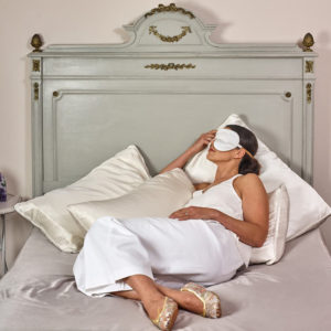 Eine Dame in einem weißen Seidenhemd liegt auf einem luxuriösen Bett mit ebenfalls weißt Satinbettwäsche und trägt eine weiße Schlafmaske von Holistic Silk