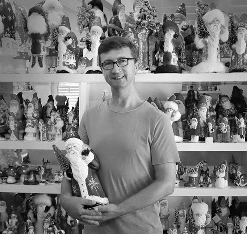 Schwarz-Weiß Foto von Thomas Schaller der Marke Ino Schaller, der vor einem Regal mit seinen Nikoläusen aus Papiermaché steht, während er einen davon in der Hand hält