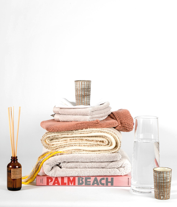 Auf dem Coffee Table Book "Palm Beach" von Assouline liegen mehrere zusammengefaltete Handtücher verschiedener Marken des RAUM concept stores - davor steht ein Diffuser von P.F. Candles, eine gefüllte Wasserkaraffe, sowie ein Keramikbecher