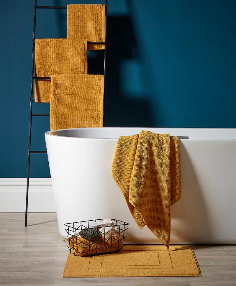 Safranfarbene Handtücher von Christy auf einem Handtuchhalter mit einem safranfarbenen Badvorleger vor einer Badewanne